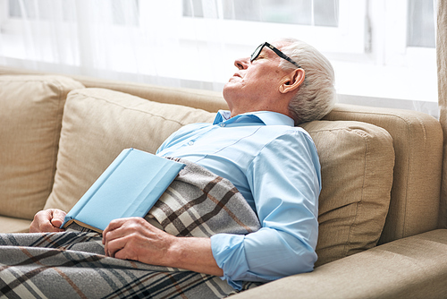 Tired senior man covered with plaid napping on couch with open book that he did not finishand fell asleep while staying at home