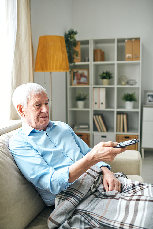 Content relaxed elderly man with blanket on legs sitting on sofa and using remote control while watching tv alone at home