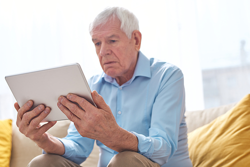 Below view of serious elderly man in shirt sitting on sofa and watching movie with interest on tablet
