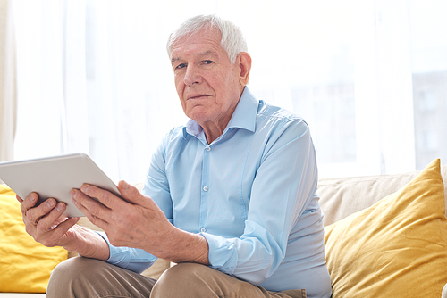 Serious senior man in casualwear looking at you while sitting on couch and using digital tablet in home environment