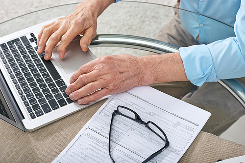 Close-up of unrecognizable senior man sitting at glassy table and using laptop while filling claim form online