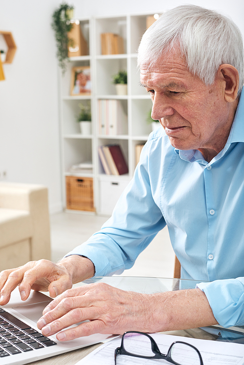 Aged retired man in blue shirt pressing keys of laptop keypad while surfing in the net for online data in home environment