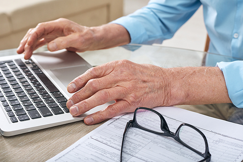 Close-up of unrecognizable elderly man sitting at table and using online service on laptop