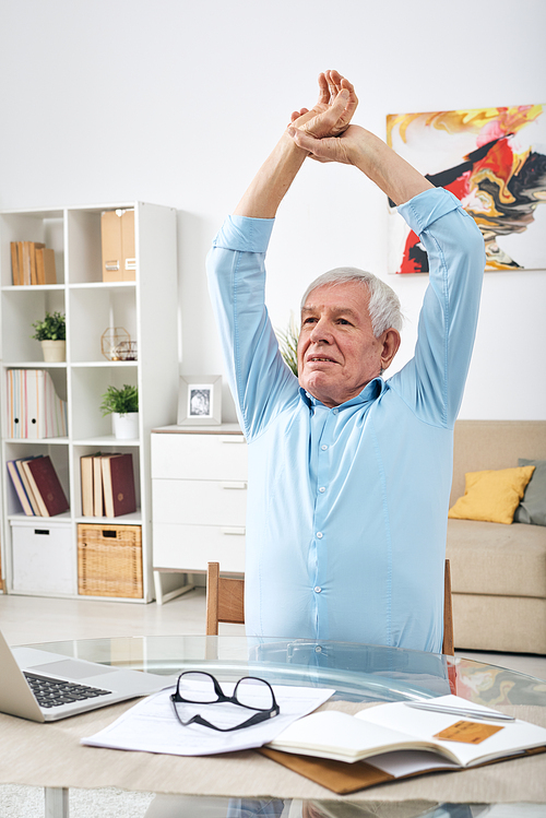 Senior slightly tired man raising his hands over head while exercising by table in front of laptop during work at home