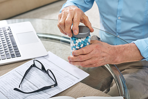 Close-up of unrecognizable senior man sitting at table with laptop and opening pill jar while preparing to take medication