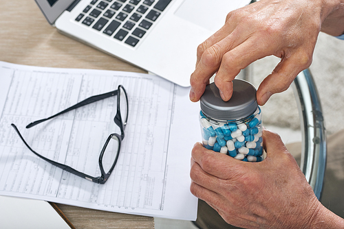 Hands of sick senior man uncovering jar with pills to take medicine while sitting by table during work with documents