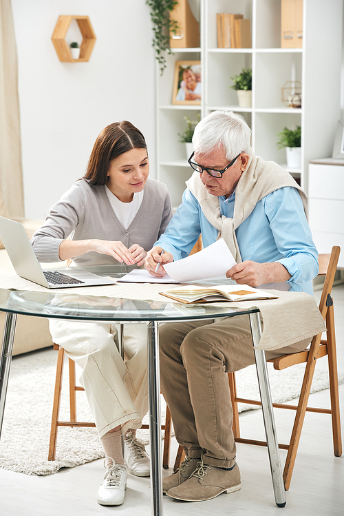 Young insurance agent sitting at table in living room and assisting senior man to fill papers
