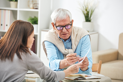 Young social worker or insurance agent asking questions of senior man about points of the paper during home consultation