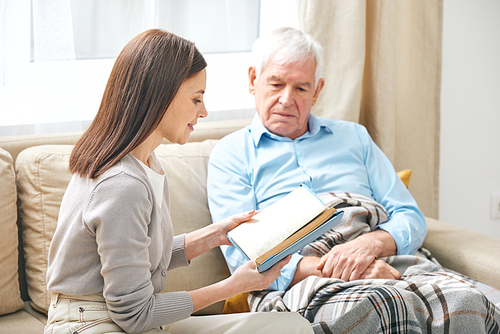 Smiling young female social worker sitting on sofa and reading book to elderly man under blanket