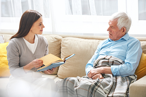 Young female social worker with open book discussing its subject with senior grey-haired man while both sitting on couch