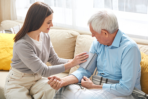 Young nurse sitting on sofa and taking pressure of elderly man in living room, she visiting patient at home