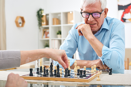 Unrecognizable woman sitting at table and playing chess with retired senior man in glasses in nursing house