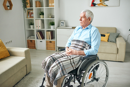 Sad and serene senior disable man with checkered plaid on his knees sitting in wheelchair by couch at home