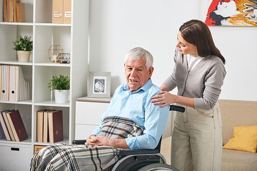 Careful young nurse talking to elderly man in wheelchair while visiting him at home