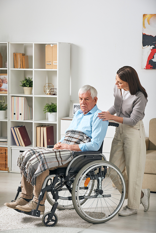 Young female care giver talking to grey-haired senior male pensioner sitting in wheelchair while staying at home