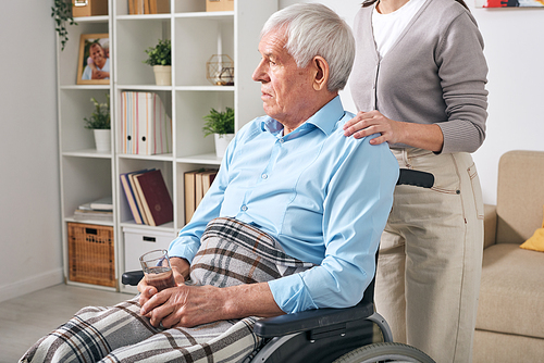 Aged disable man with glass of water sitting on wheelchair with young female caregiver standing behind and comforting him