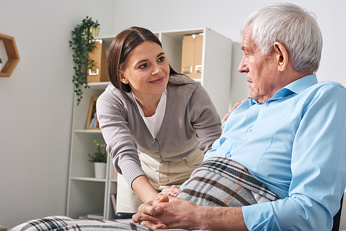 Friendly young female nursing care specialist asking senior man about his needs while looking after him