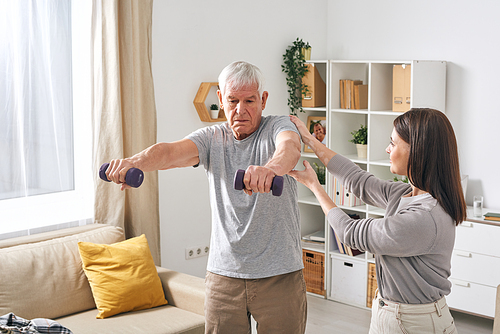 Young female social worker helping aged man in activewear exercising with dumbbells in home environment