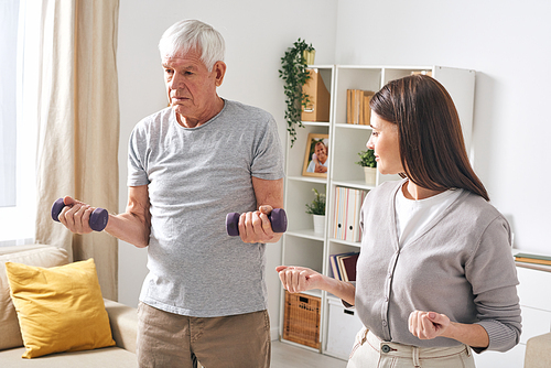 Young social worker showing senior man how to perform exercise with dumbbells while helping him to rehab