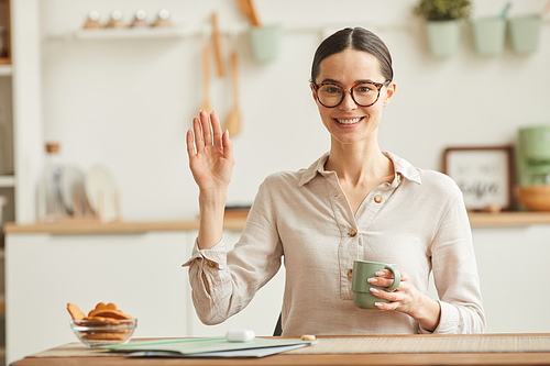 Warm toned portrait of young woman wearing glasses and waving at camera while sitting at desk in cozy home interior, copy space
