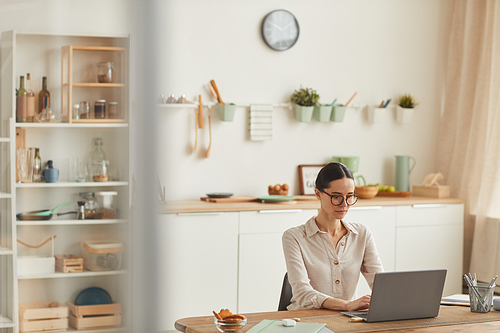 Warm-toned wide angle portrait of elegant young woman wearing glasses while using laptop at cozy home office workplace, copy space