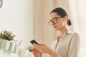 Warm-toned portrait of elegant businesswoman using smartphone while standing in cozy home interior, copy space