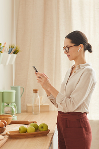 Vertical warm-toned portrait of businesswoman using smartphone while standing in cozy home interior, copy space