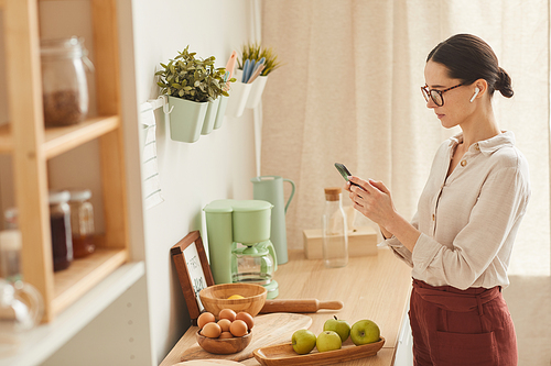 Side view portrait of elegant modern woman holding smartphone and using wireless earphones while standing in cozy kitchen interior, copy space