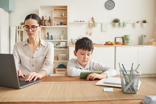 Warm toned portrait of young mother working at home office with son using smartphone beside her in cozy kitchen interior, copy space
