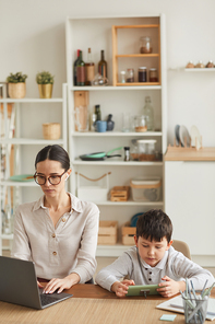 Vertical portrait of young mother working at home office with son using smartphone beside her in cozy kitchen interior, copy space