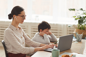 Warm toned side view portrait of young mother working at home office with son using smartphone beside her in cozy kitchen interior, copy space