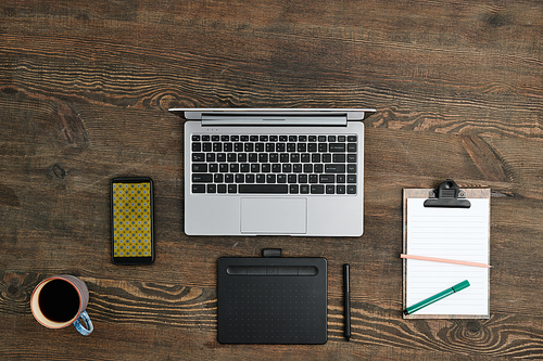 Flat layout of graphic tablet with stylus, mobile gadgets, mug with tea or coffee and clipboard with working document on wooden table