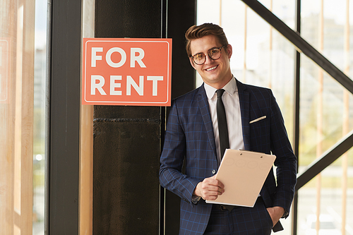Waist up portrait of smiling real estate agent  while posing next to red FOR RENT sign indoors, copy space