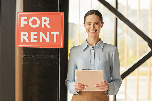 Waist up portrait of female real estate agent smiling happily  while posing next to red FOR RENT sign indoors, copy space
