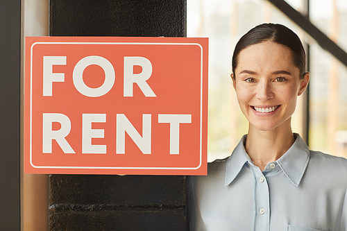 Head and shoulders portrait of female real estate agent smiling happily  while posing next to red FOR RENT sign, copy space
