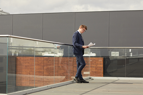 Wide angle view of young successful businessman reading documents while standing on roof of modern office building, copy space