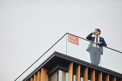 Wide angle view at smiling real estate agent speaking by phone while standing on roof of modern building next to red FOR RENT sign, copy space