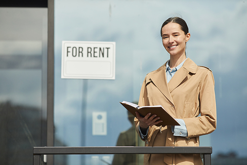 Waist up portrait of female real estate agent smiling happily  while standing next to FOR RENT sign outdoors, copy space