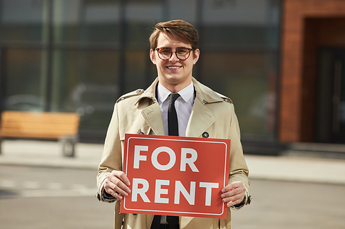 Waist up portrait of male real estate agent smiling happily  while holding red FOR RENT sign outdoors, copy space