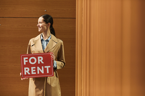 Waist up portrait of female real estate agent smiling happily looking away while holding red FOR RENT sign against graphic wooden background, copy space