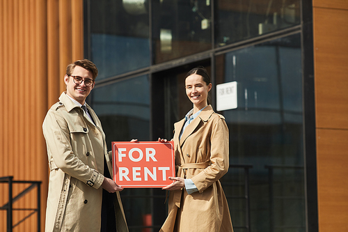 Portrait of two real estate agents holding red FOR RENT sign and smiling at camera while standing outdoors against modern office building, copy space