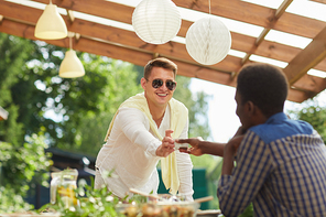 Portrait of smiling young man wearing sunglasses passing sauce to friend across table while enjoying outdoor dinner at Summer party, copy space