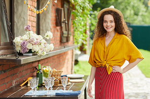Waist up portrait of young curly haired woman smiling at camera while posing by table at outdoor party in Summer, copy space