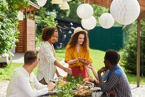 Multi-ethnic group of people enjoying dinner at outdoor terrace in Summer, focus on smiling young woman handing fresh fruits and berries across table, copy space