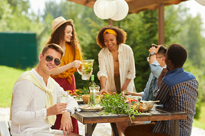 Multi-ethnic group of friends enjoying dinner at outdoor terrace in Summer, focus on young man wearing sunglasses smiling at camera, copy space