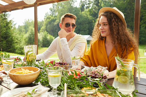 Colorful portrait of young couple enjoying dinner with friends while sitting at table during outdoor Summer party, copy space