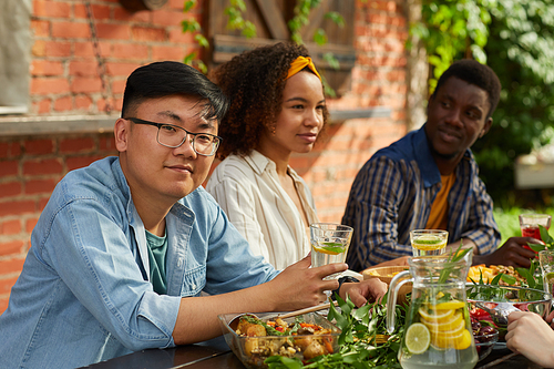 Portrait of smiling Asian man enjoying dinner with friends outdoors and  while sitting at table during Summer party, copy space