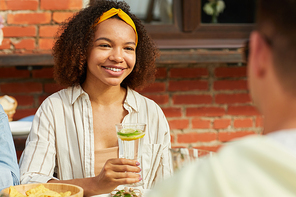 Portrait of smiling African-American woman enjoying dinner with friends outdoors and holding refreshing cocktail while sitting at table during Summer party, copy space