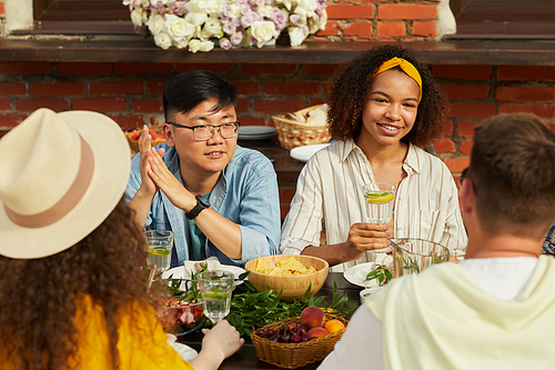 Portrait of young people enjoying dinner with friends outdoors and holding refreshing cocktails while sitting at table during Summer party, copy space