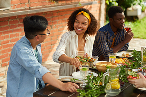 Portrait of smiling African-American woman holding potato dish while enjoying dinner with friends and family outdoors at Summer party, copy space